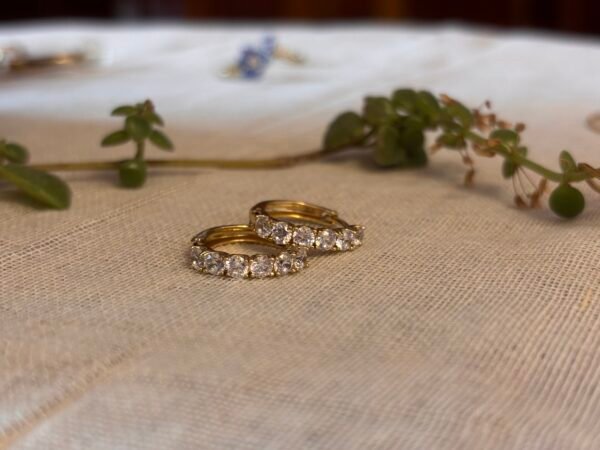 Loona Jewellery's gold huggies featuring round-cut crystals, displayed on a textured beige fabric with natural greenery in the background.